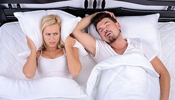 Woman covering her ears while lying in bed next to a man with his mouth open